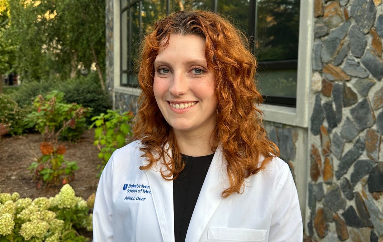 Smiling young woman with curly red hair wearing a white lab coat with 'Duke University School of Medicine' embroidered on it, standing outside by a building with large windows and stone facade.